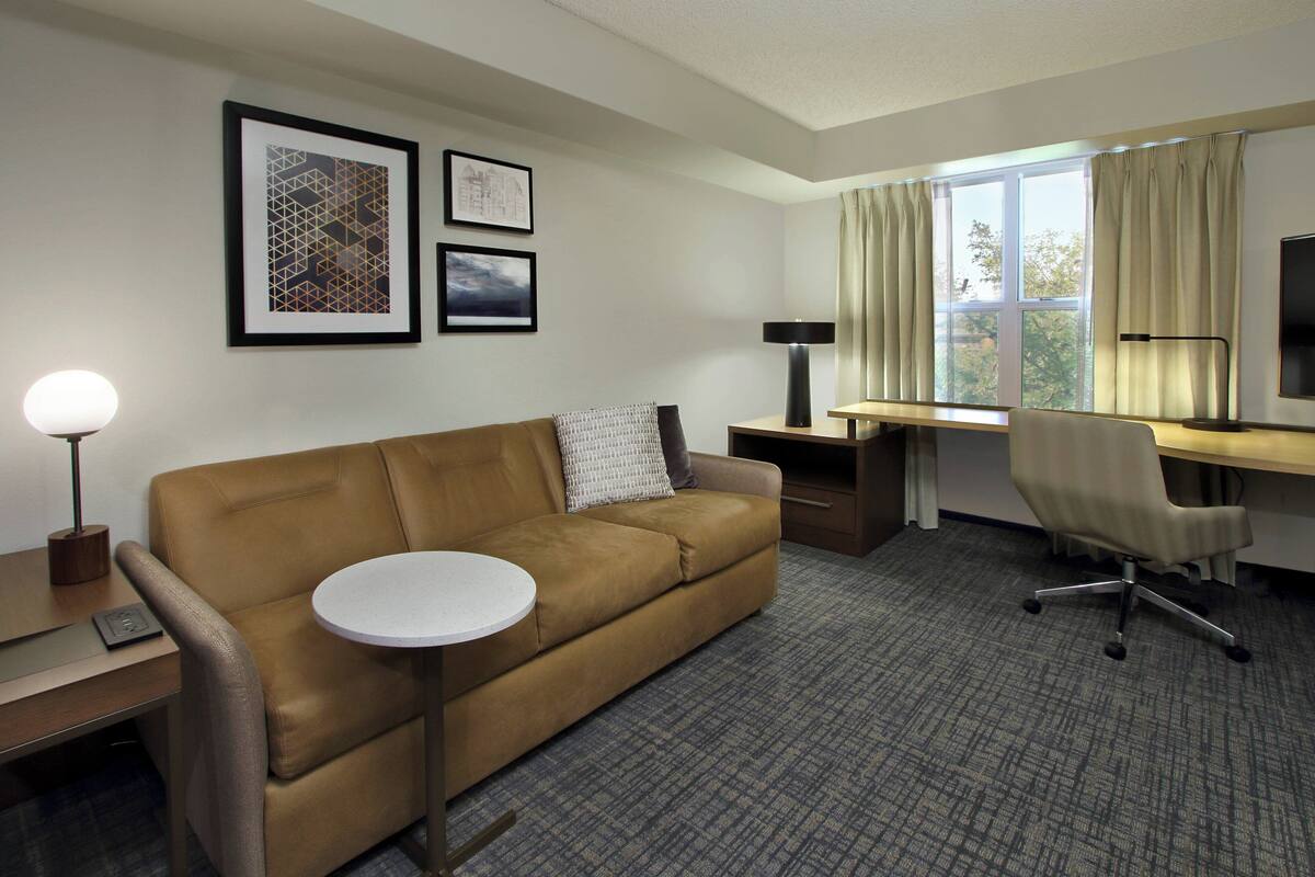 A hotel room with a brown sofa, a round table, a desk, a chair, and a TV. There are framed pictures on the wall and a window with curtains.