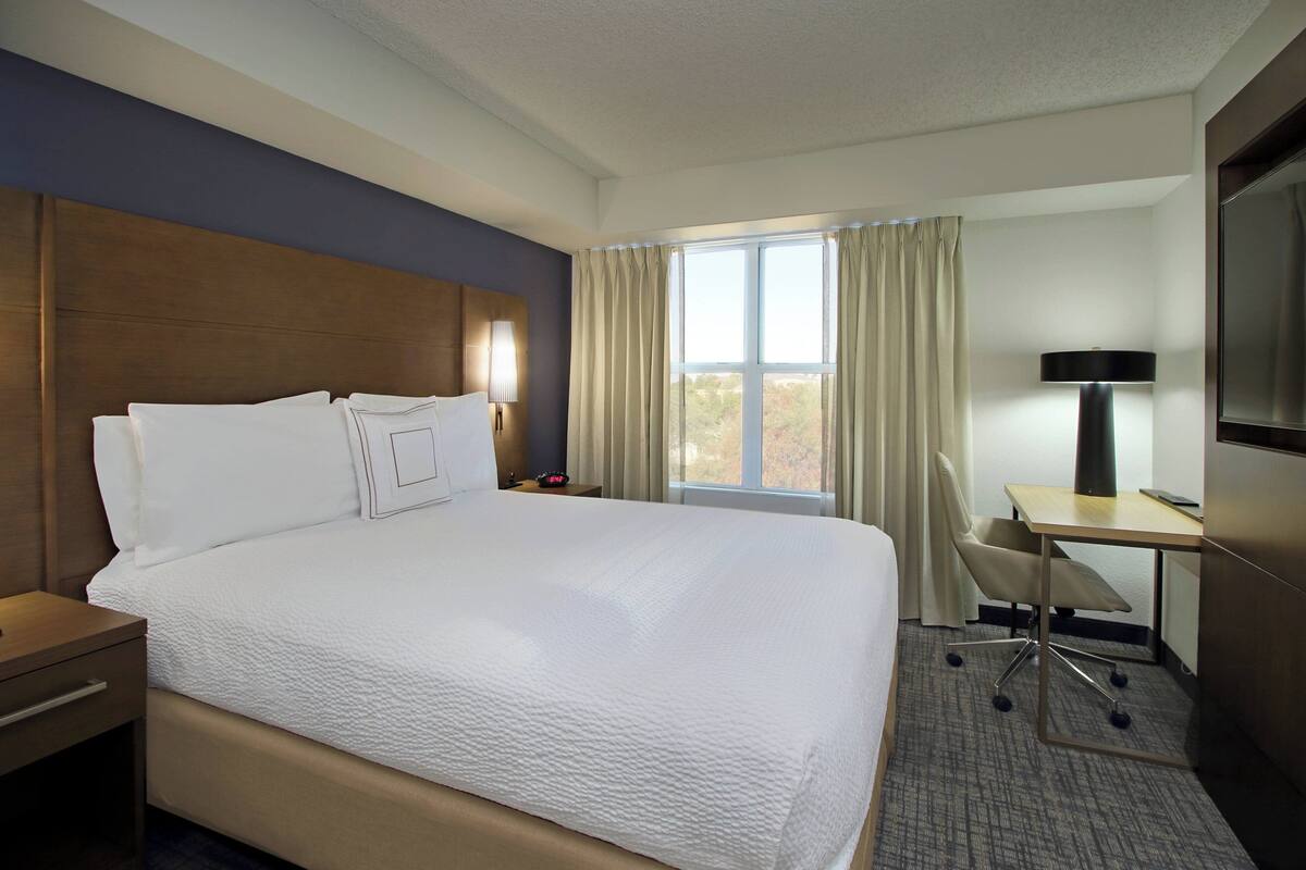 A hotel room with a large bed, a desk, a chair, a lamp, and a TV.