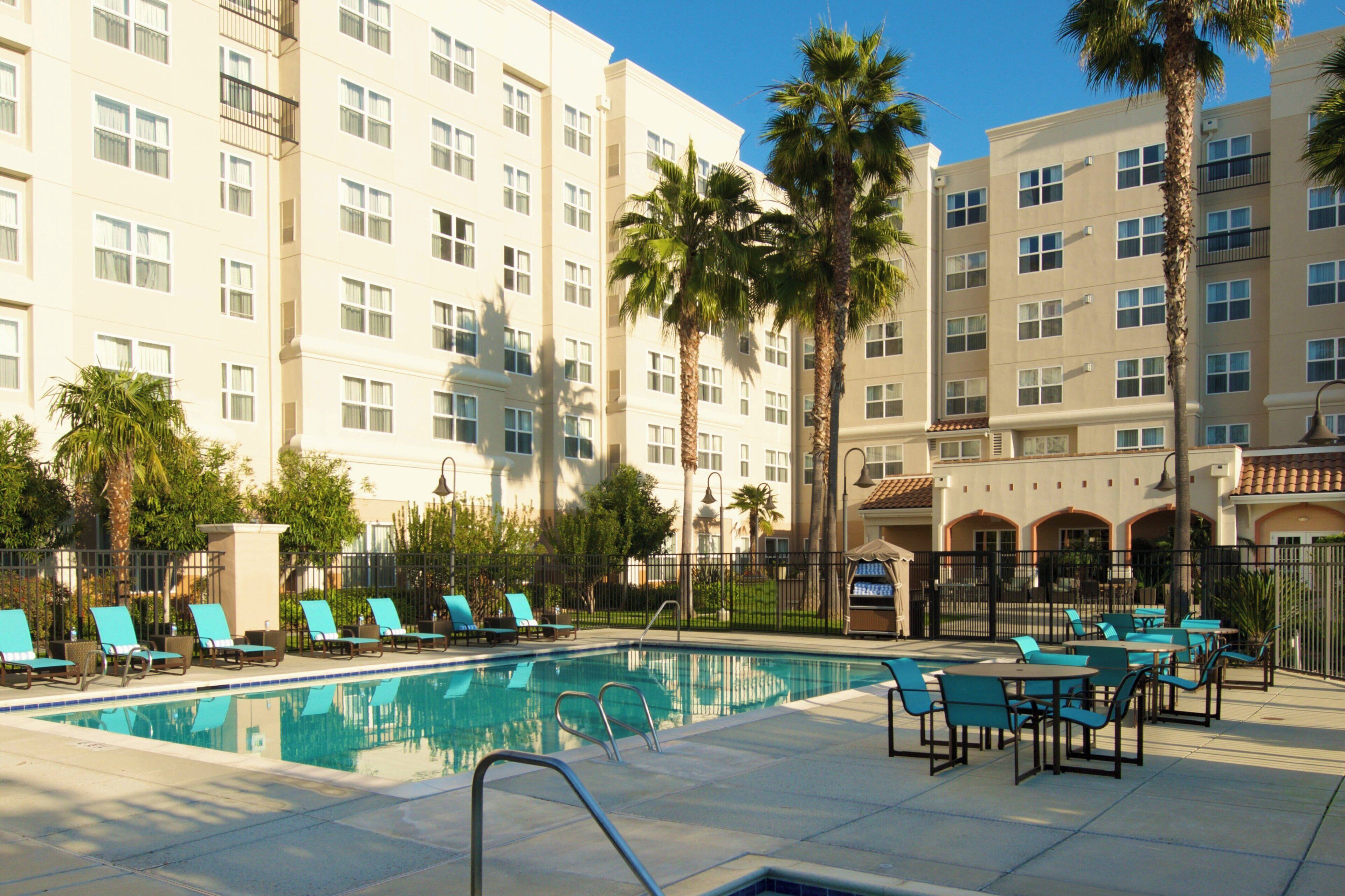A swimming pool with lounge chairs and tables, surrounded by tall buildings with balconies and palm trees.