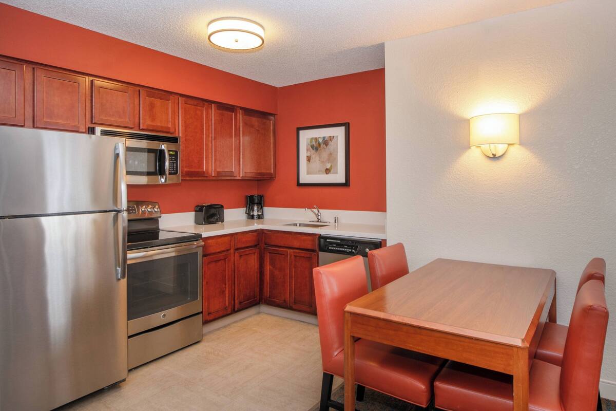 Una cocina compacta con armarios de madera, electrodomésticos de acero inoxidable y un comedor con mesa de madera y sillas rojas.