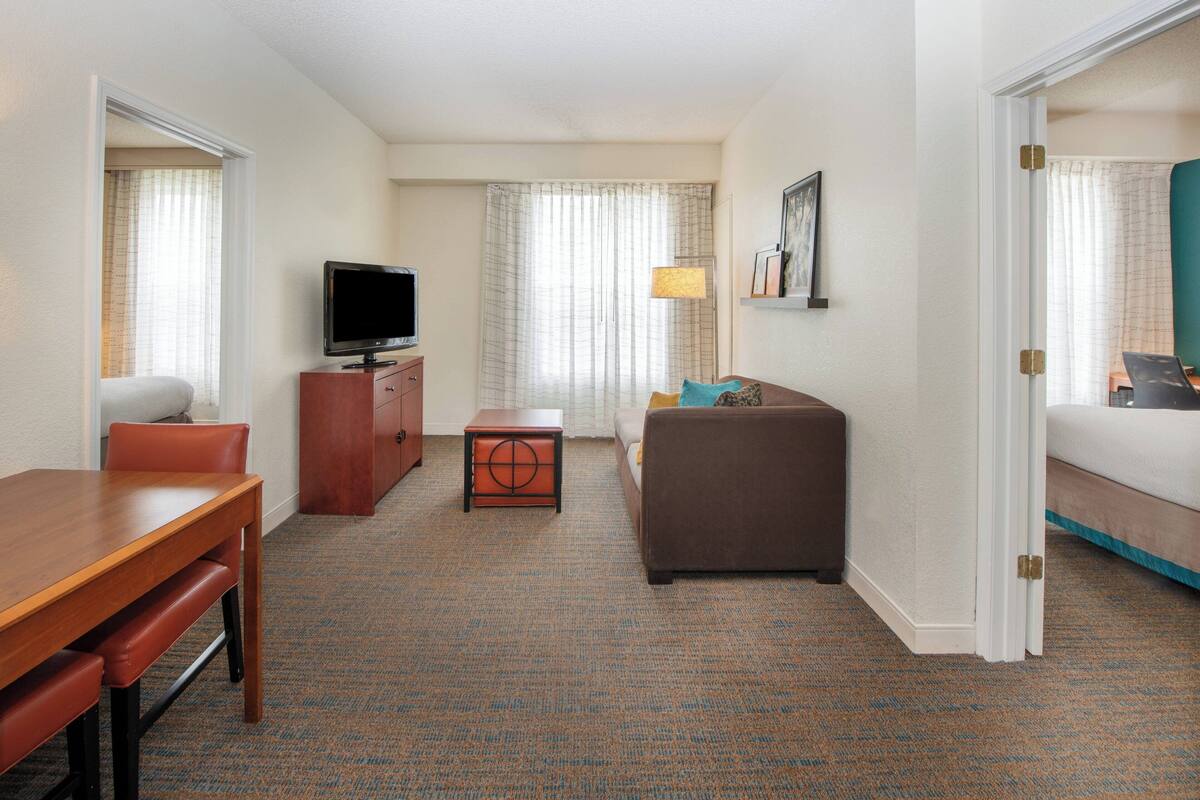 Una habitación de hotel con una mesa de comedor de madera, un sofá, un televisor sobre un mueble de madera y una ventana con cortinas.