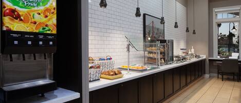 Uma mesa de pequeno-almoço com dispensador de sumo, uma vitrine de vidro e diversas opções de comida.