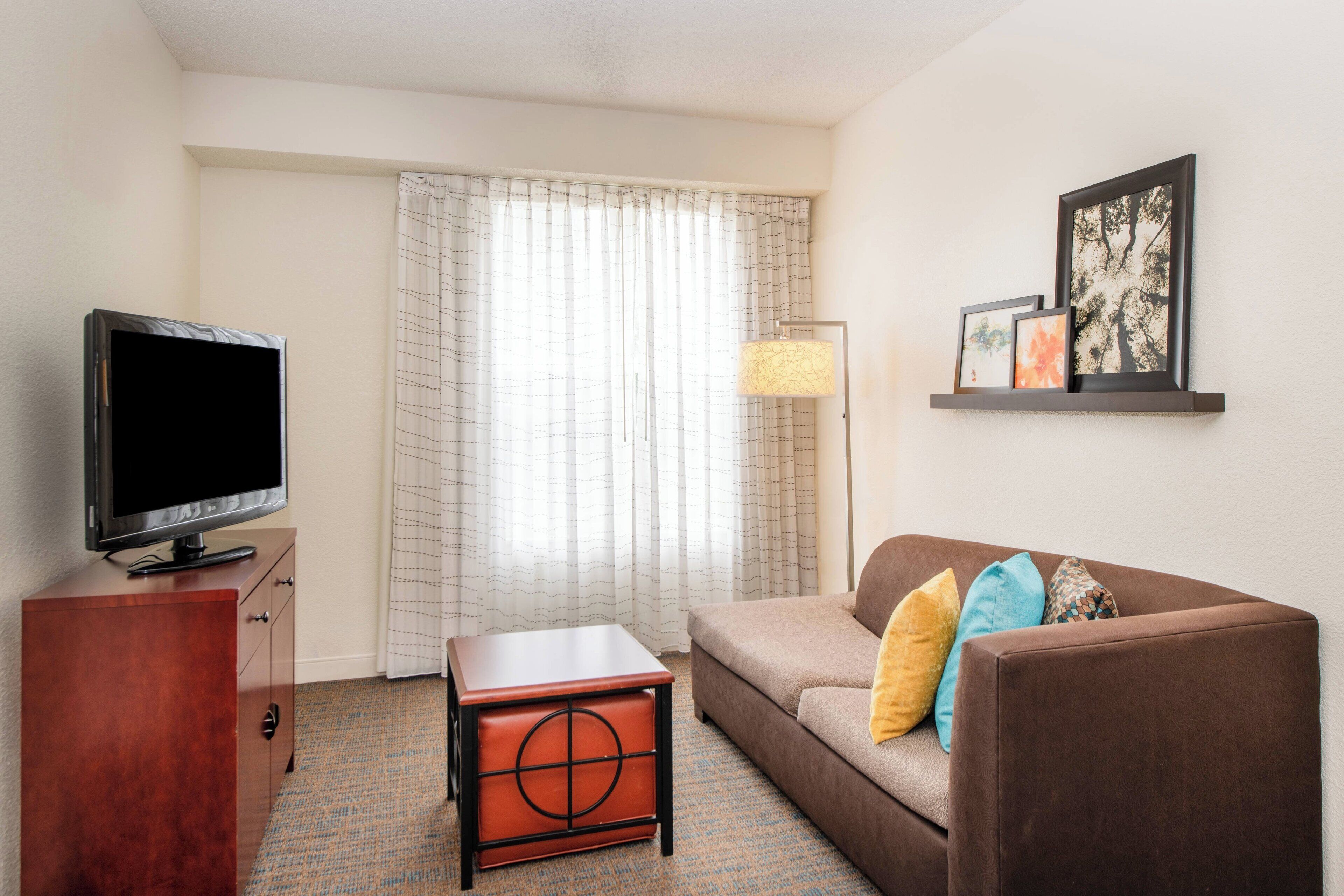 A hotel room with a flat-screen TV on a wooden cabinet, a brown sofa with colorful cushions, a small wooden side table, a floor lamp, and a large window with sheer curtains.