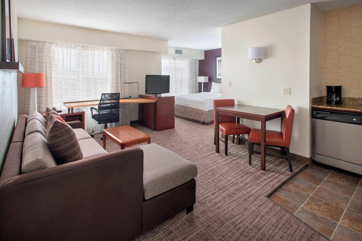 Habitación de hotel con sofá, una mesita, escritorio con silla, cama, televisión y área de cocina.
