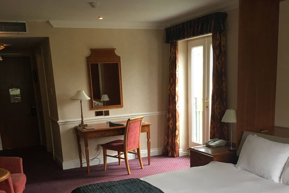 A hotel room with a large bed, a desk with a chair, a small table with a lamp, and a window with curtains.