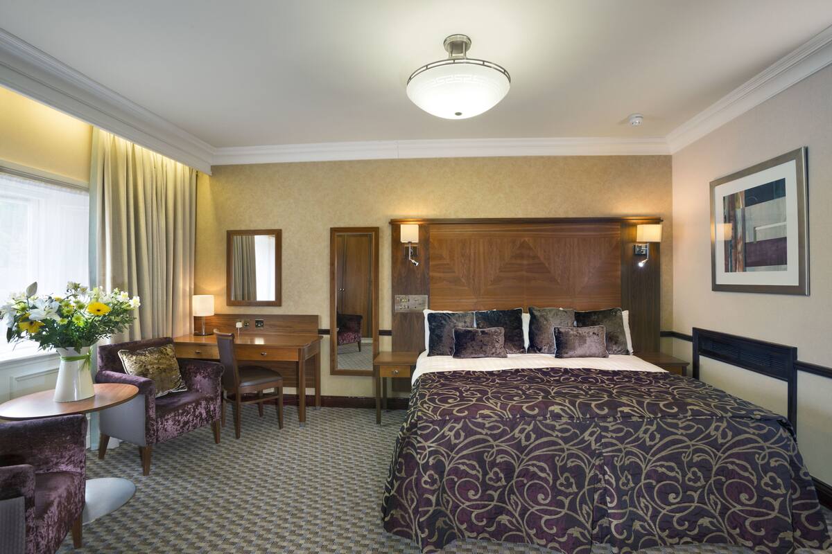 A hotel room with a large bed, a desk with a chair, a small table with a vase of flowers, and a mirror.
