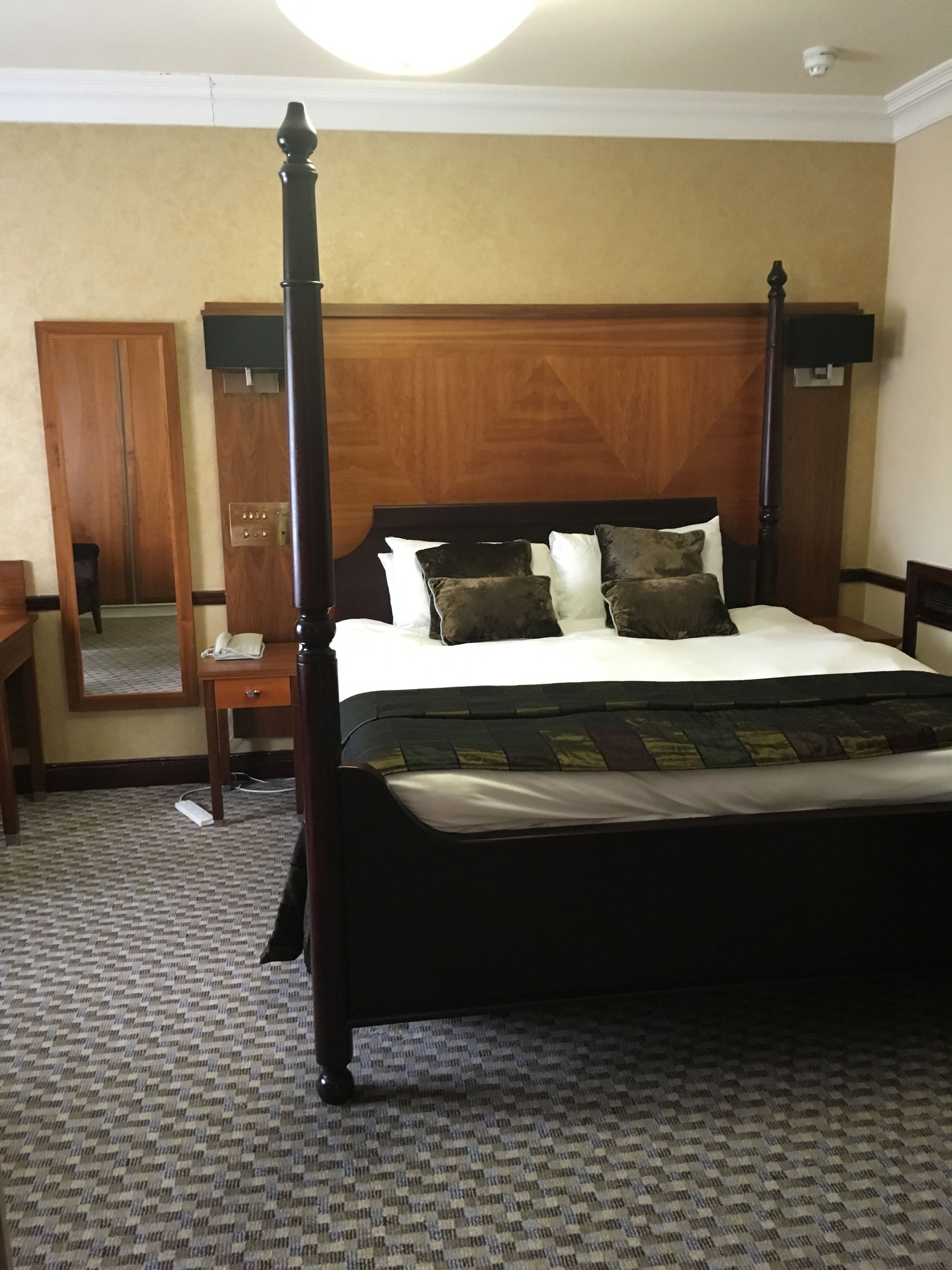 A four-poster bed with a wooden headboard, a bedside table with a lamp, and a mirror on the wall.
