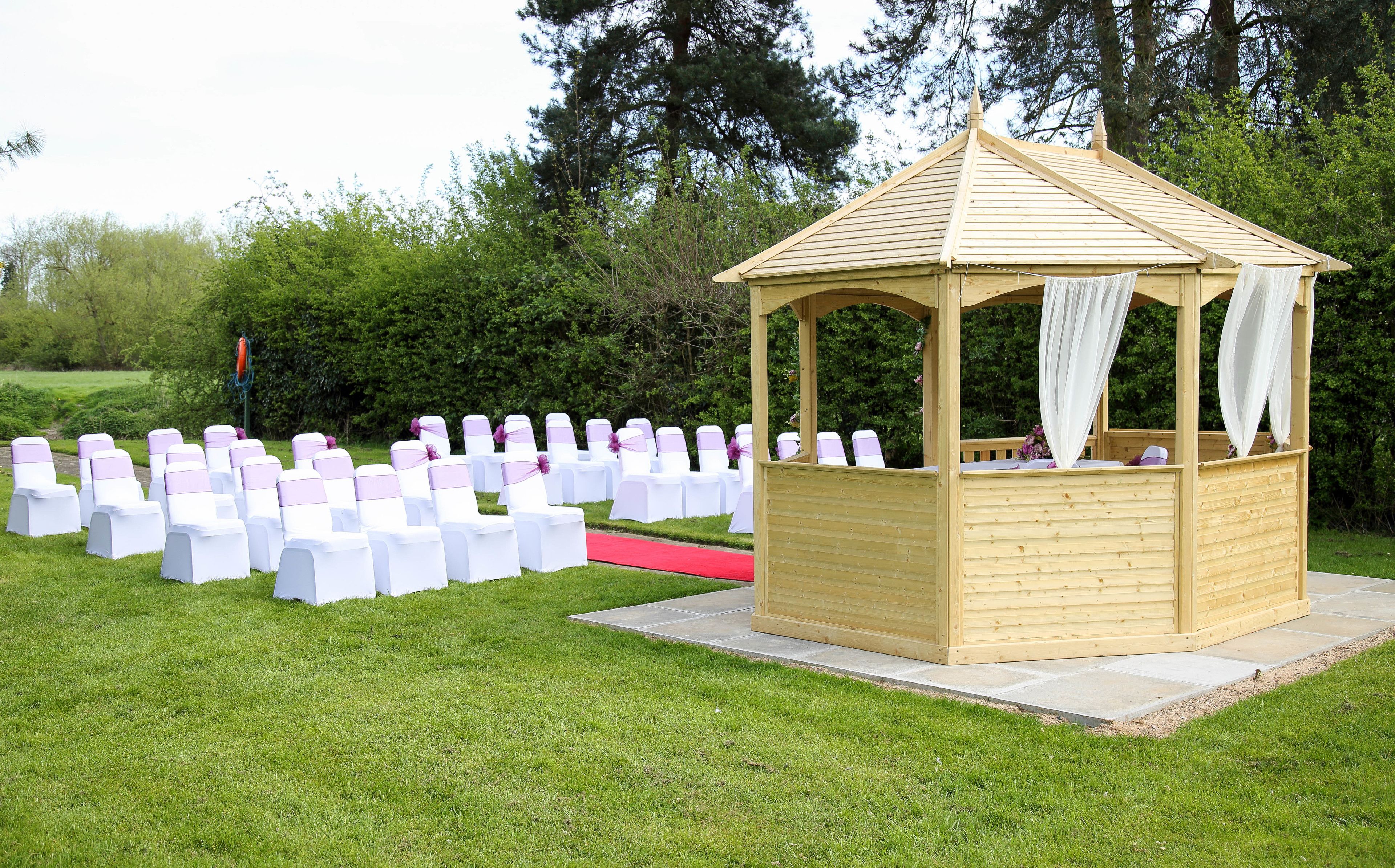 A wooden gazebo with a red carpet leading to it, surrounded by white chairs with purple sashes.