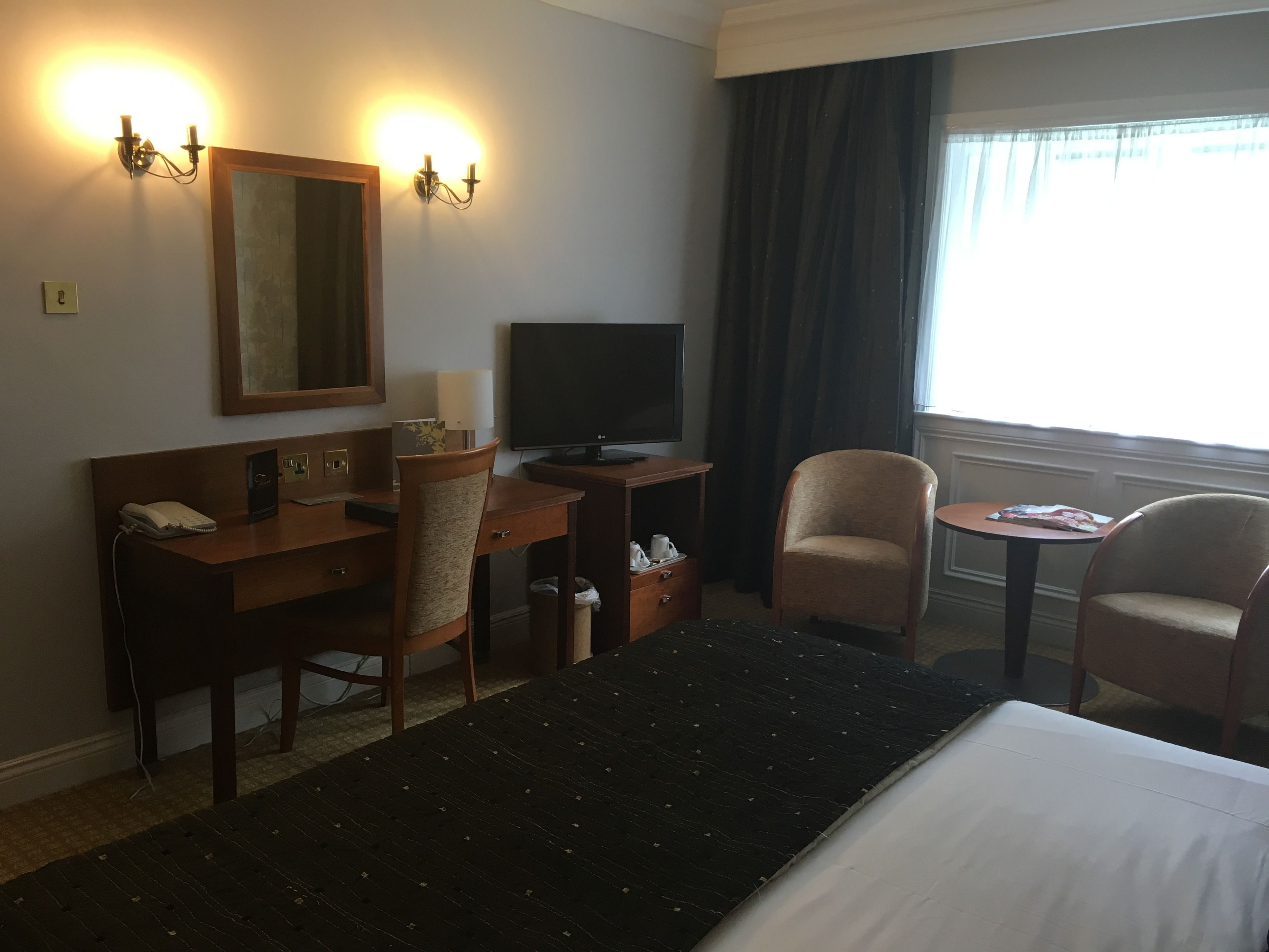 A hotel room with a bed, a desk with a telephone, a chair, a television, and a window with curtains.
