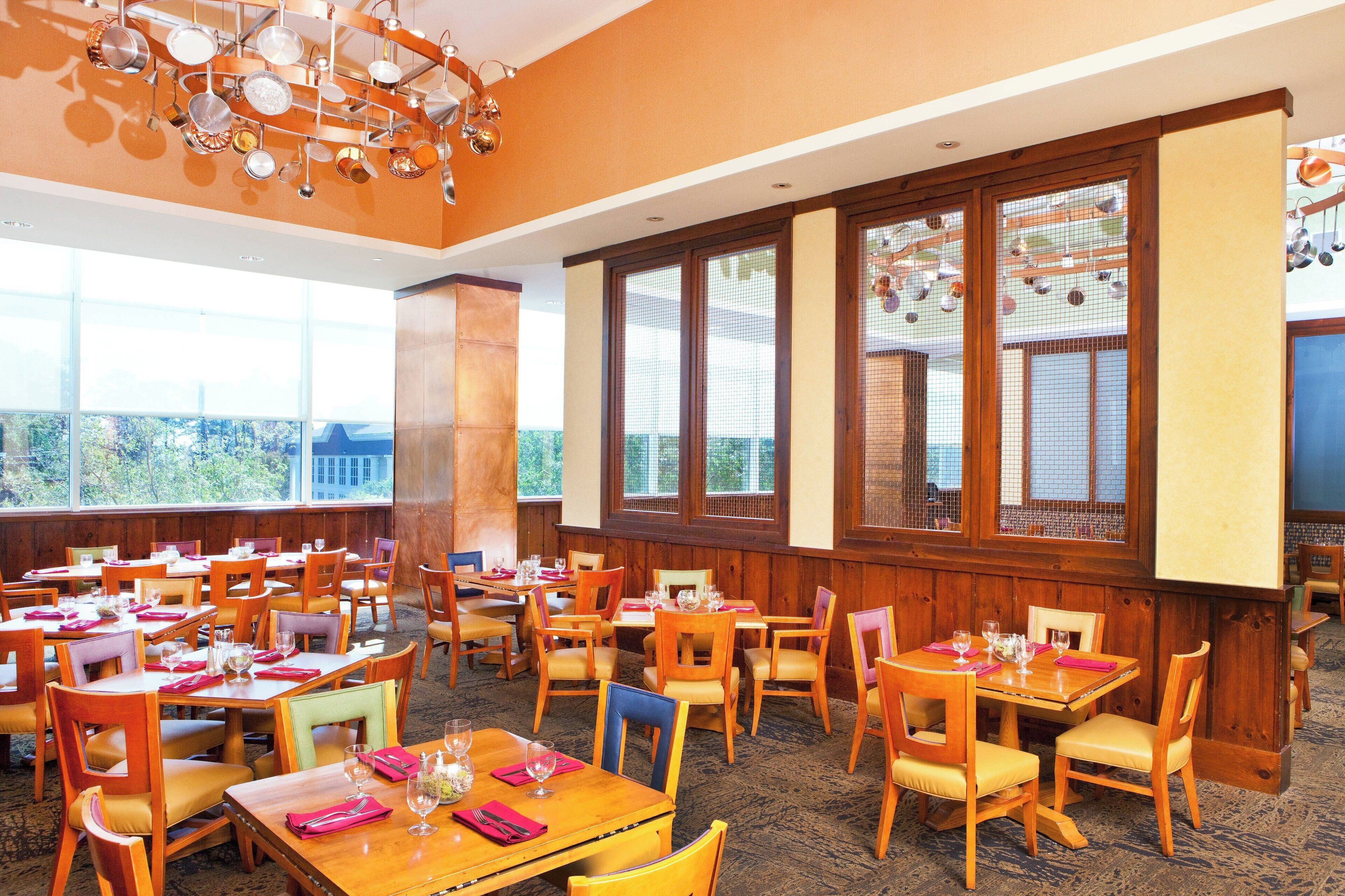 Un comedor con varias mesas y sillas, ventanas amplias y un elegante candelabro.