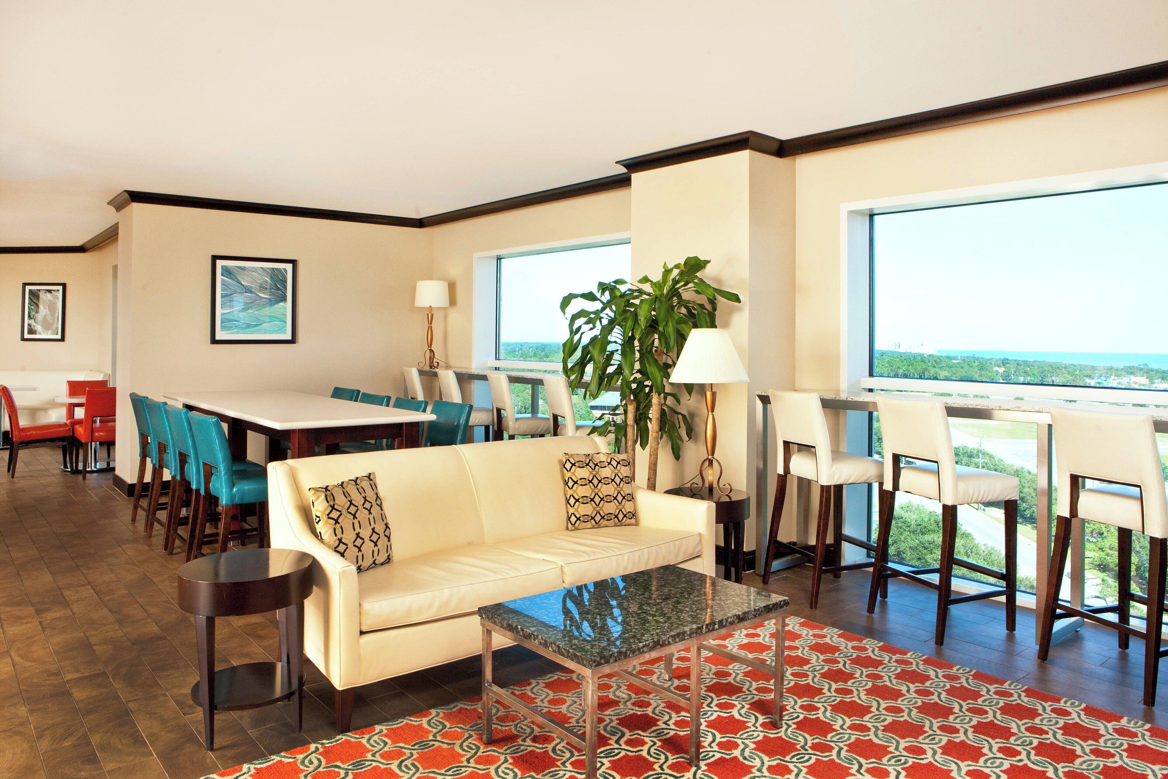 A modern hotel room with a dining area, a sofa, a coffee table, and a large window offering a view.