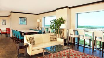 A modern hotel room with a dining area, a sofa, a coffee table, and a large window offering a view.