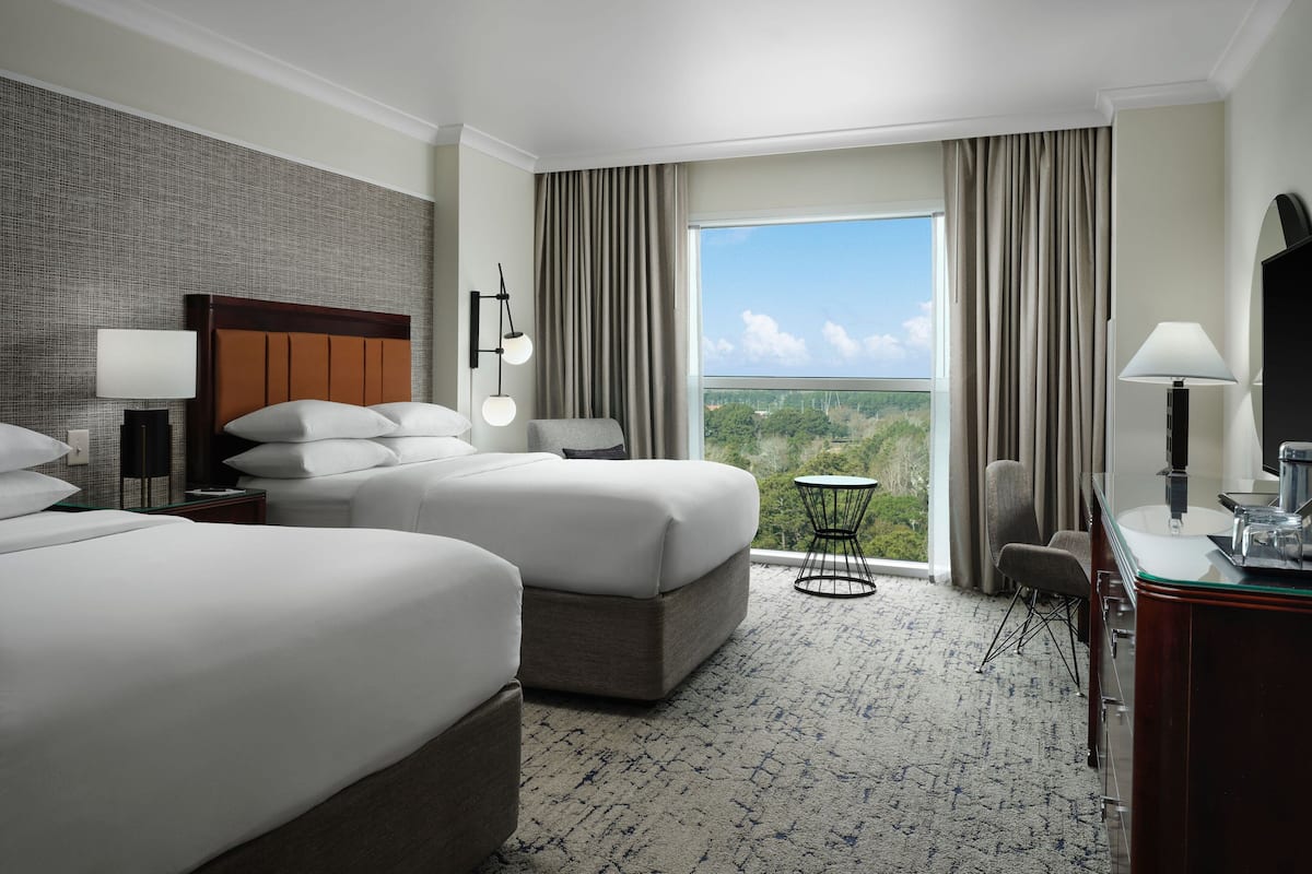 A hotel room with two beds, a desk, a chair, a small table, and a large window offering a view of the outdoors.