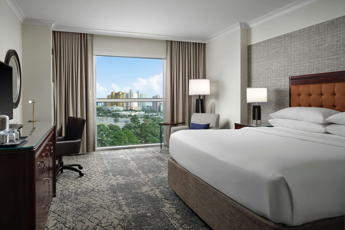 A hotel room with a large bed, a desk with a chair, a desk lamp, and a view of the cityscape through a large window.