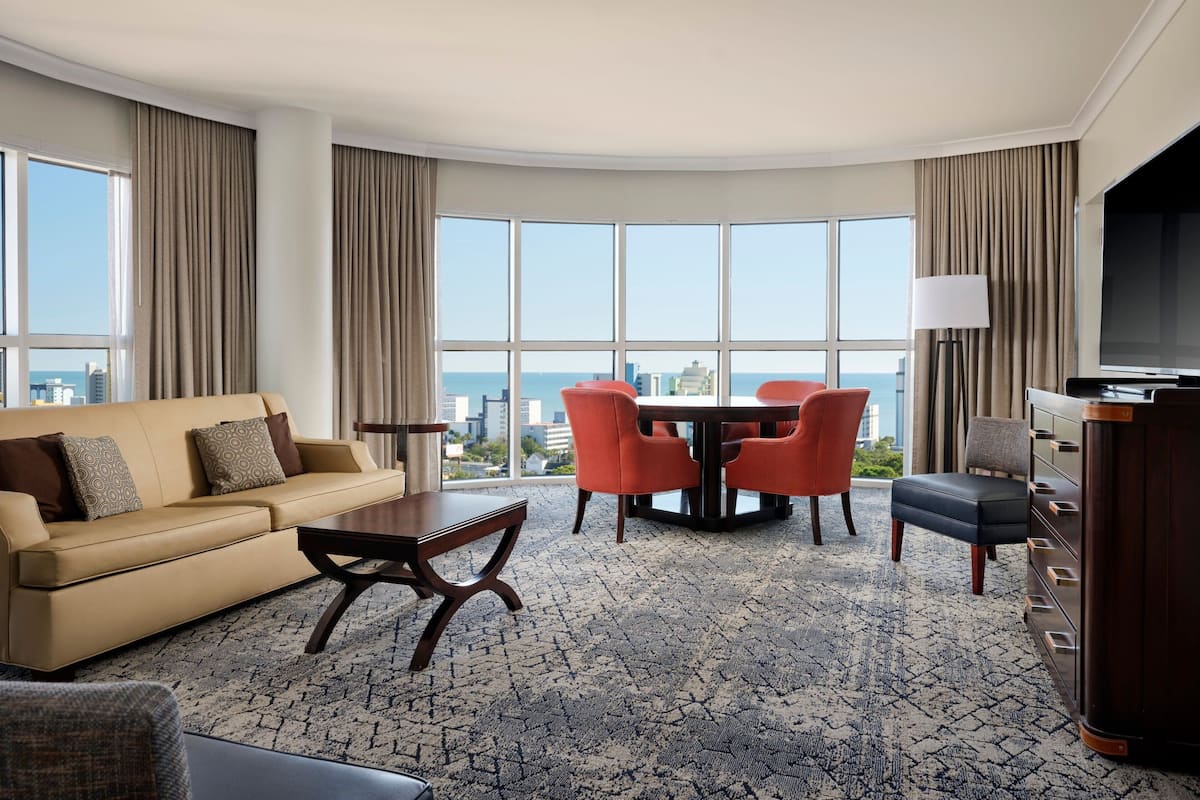 A hotel room with a large window, a sofa, a dining table with chairs, a TV, and a dresser.