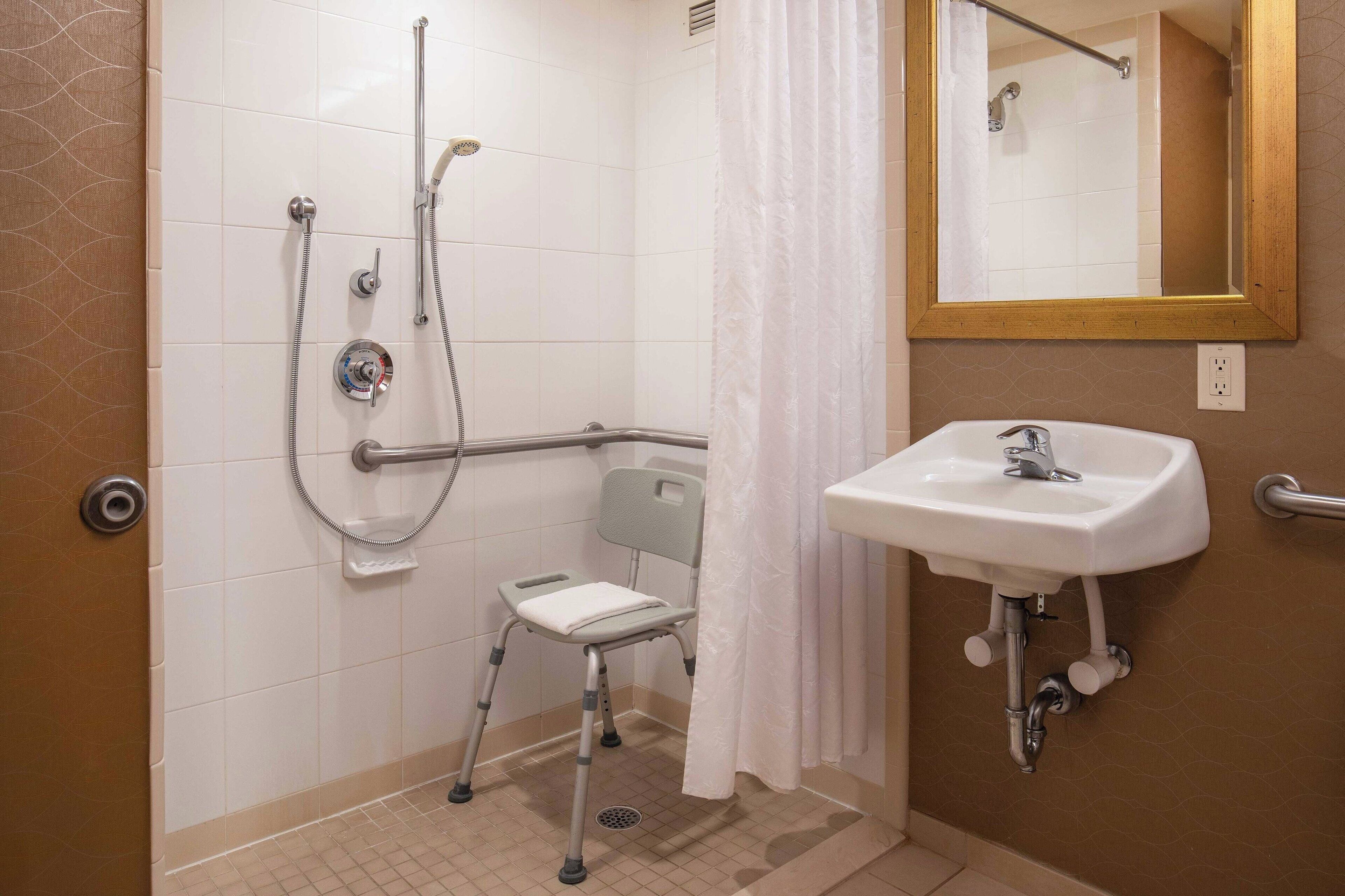 Un baño con ducha, lavamanos, espejo y silla accesible para usuarios de silla de ruedas.