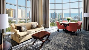 A hotel room with a large window, a beige sofa, a wooden coffee table, a small round table, and red chairs. - Sheraton Myrtle Beach (Myrtle Beach)