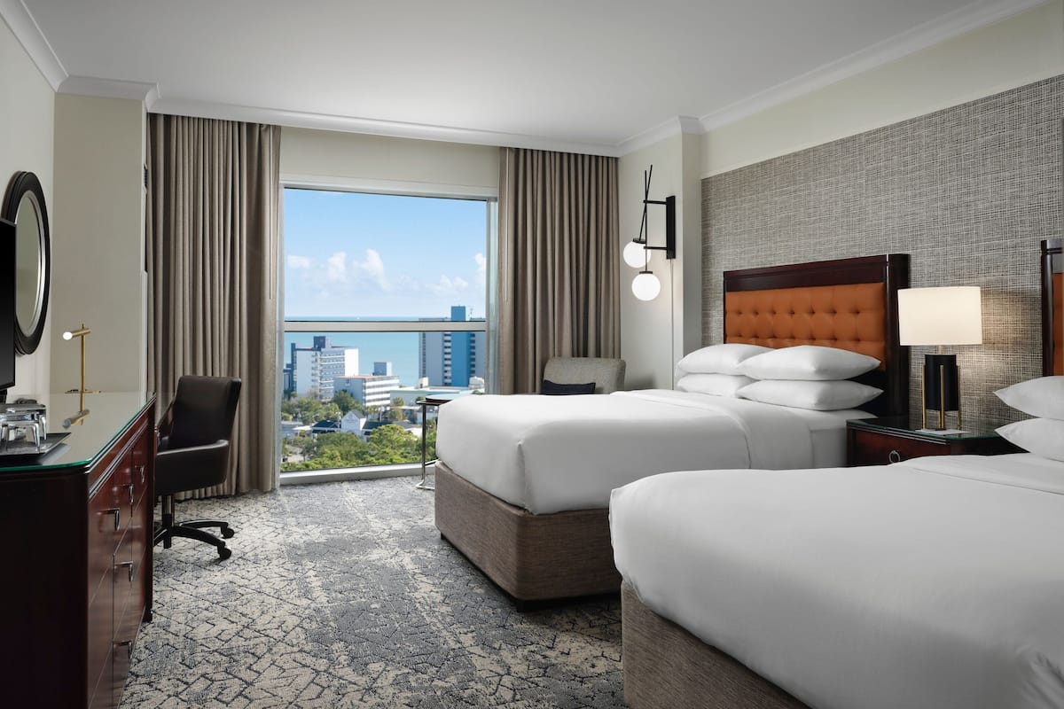 A hotel room with two beds, a desk, a chair, and a large window offering a city view.