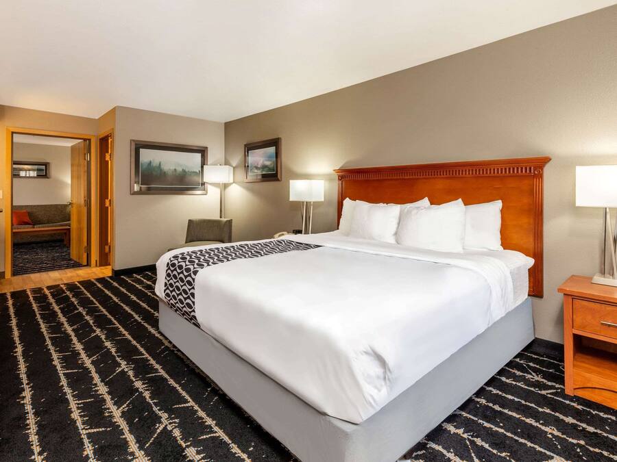 A hotel room with a large bed, a wooden headboard, two bedside tables with lamps, a black and white patterned rug, and a door leading to another room.