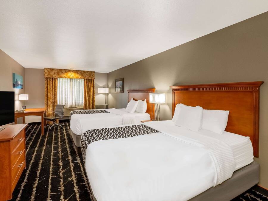 Quarto de hotel com duas camas, uma mesa, uma televisão e uma janela grande com cortinas.