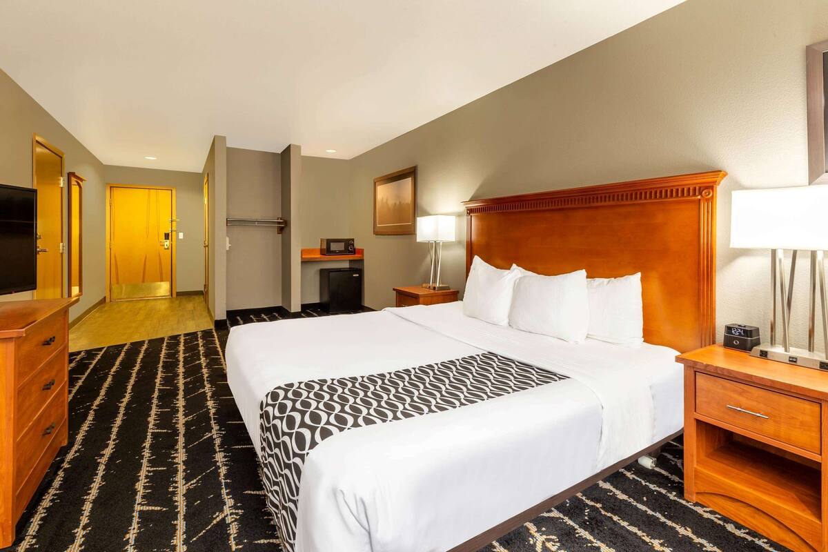 Ein Hotelzimmer mit einem großen Bett, einem Holzkopfende, einem Nachttisch mit Lampe, einem Fernseher und einem Schreibtisch mit Computer.