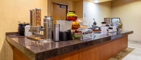 A buffet counter with various food items and containers, including a bowl of fruit and a coffee station.