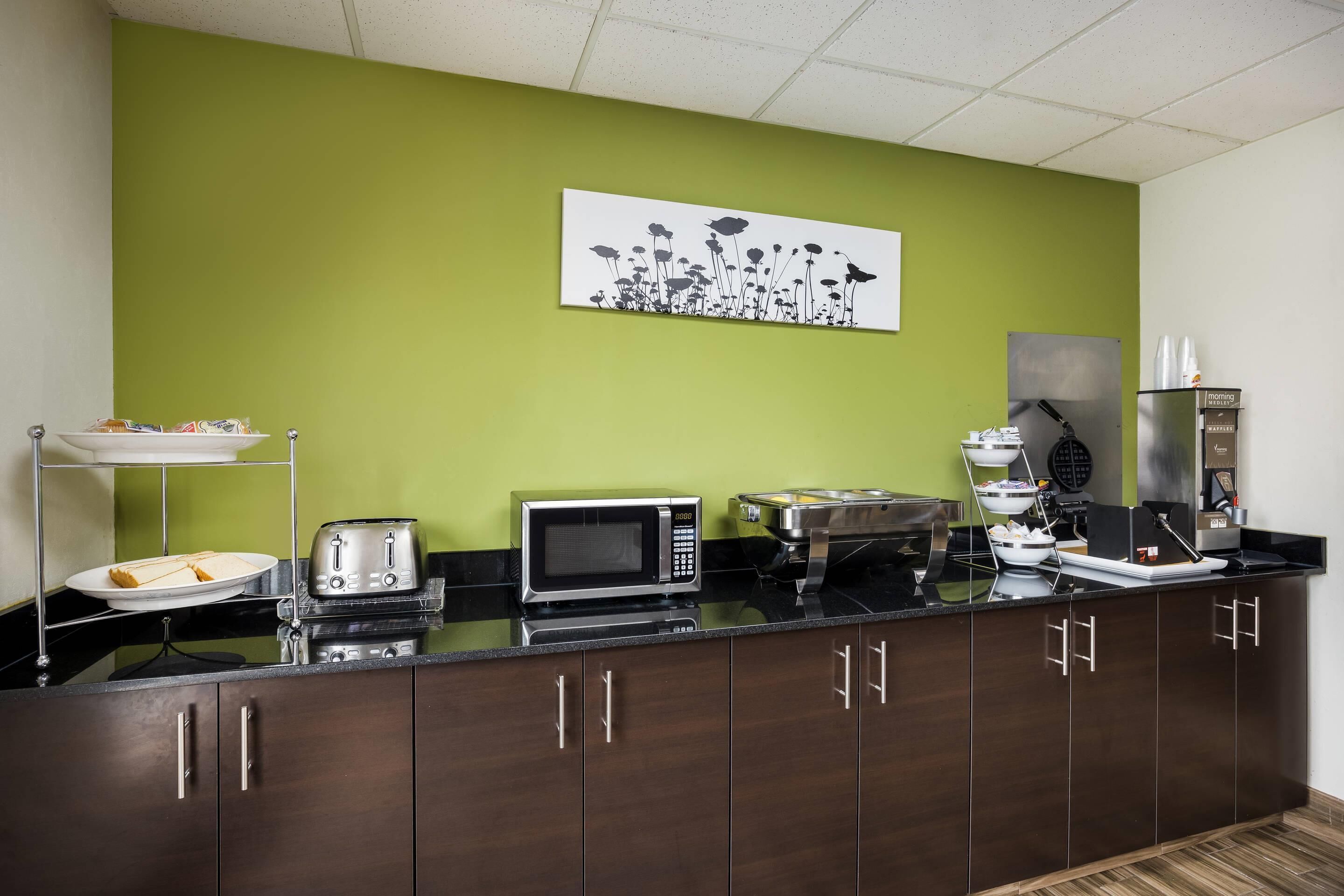 Una cocina con microondas, tostadora y cafetera sobre la encimera, y una pared verde con un cuadro floral.