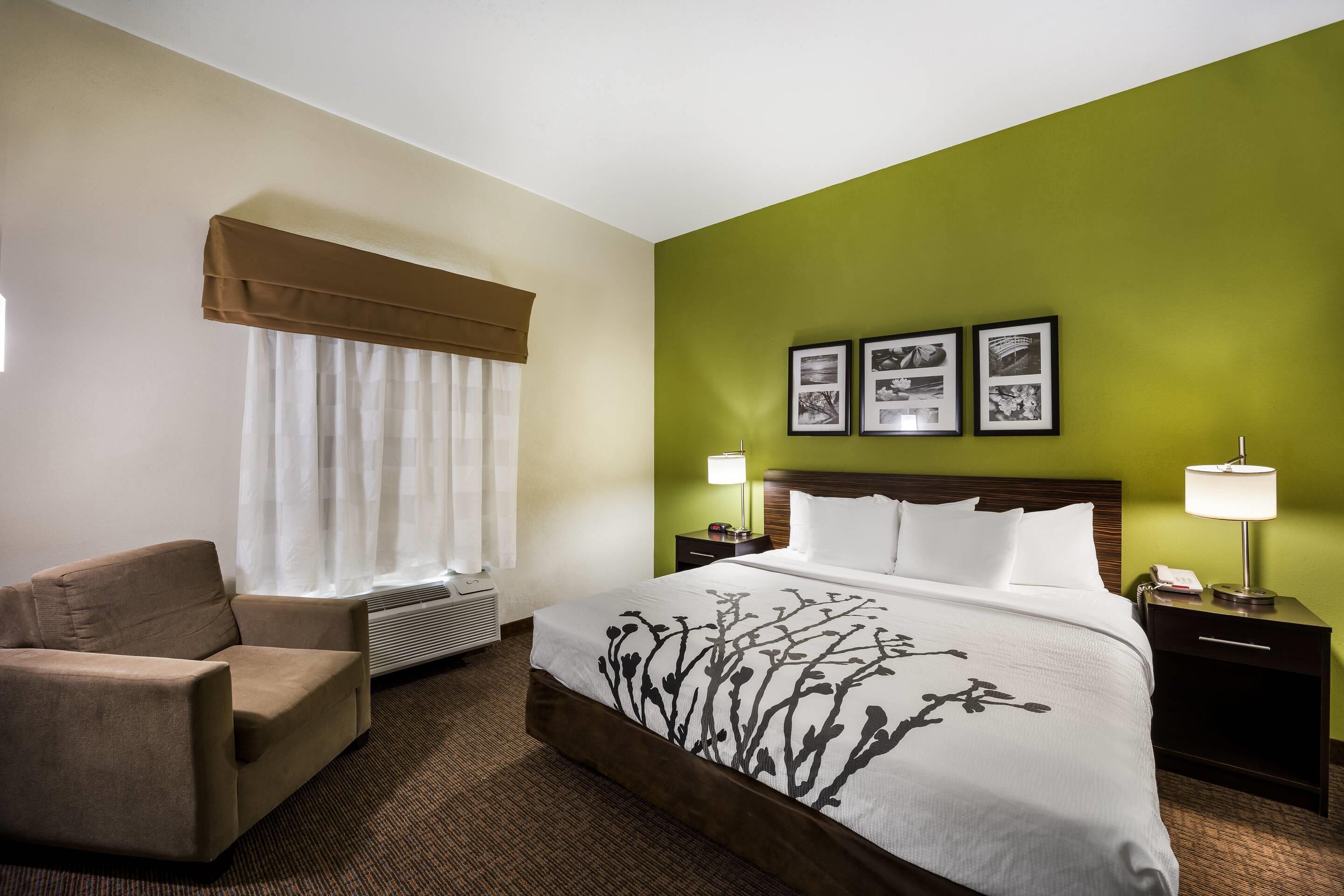 Quarto de hotel com cama, uma cadeira, mesa de cabeceira e parede com três quadros.