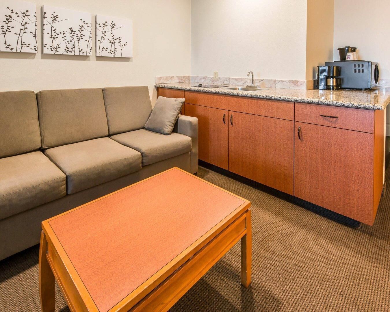 A compact living space with a sofa, a small wooden table, a kitchenette with a sink and a coffee maker, and wall-mounted artwork.