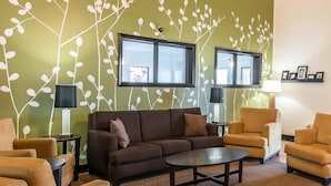 Ein Wohnzimmer mit einem braunen Sofa, zwei Sesseln, einem Couchtisch und zwei Lampen. Zwei Fenster mit Jalousien und eine Wand mit einem Blattmuster sind ebenfalls zu sehen.