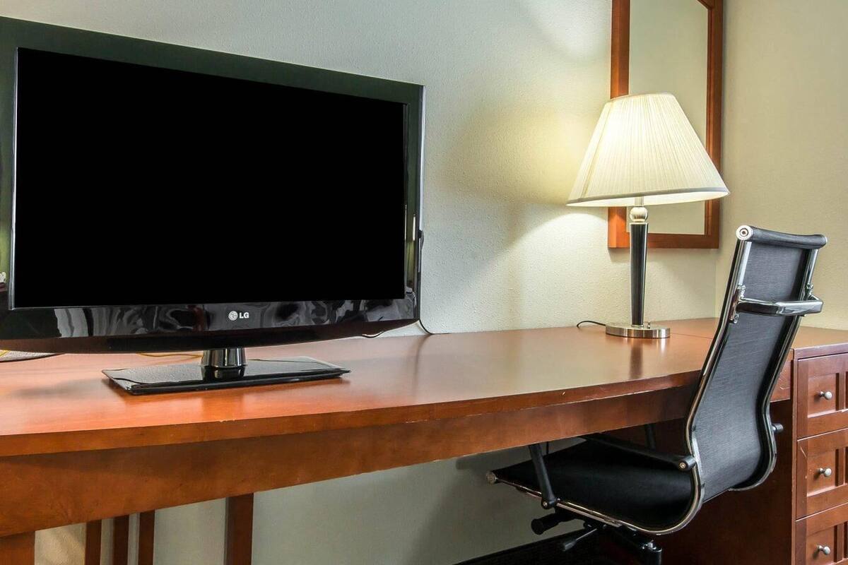 A hotel room with a wooden desk, a black office chair, a flat-screen TV, and a table lamp.