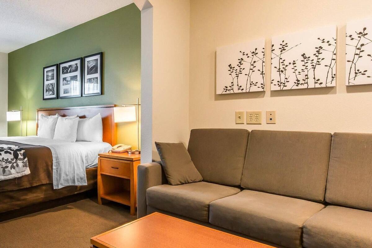 A hotel room with a bed, a sofa, a coffee table, a nightstand, a lamp, and wall art.