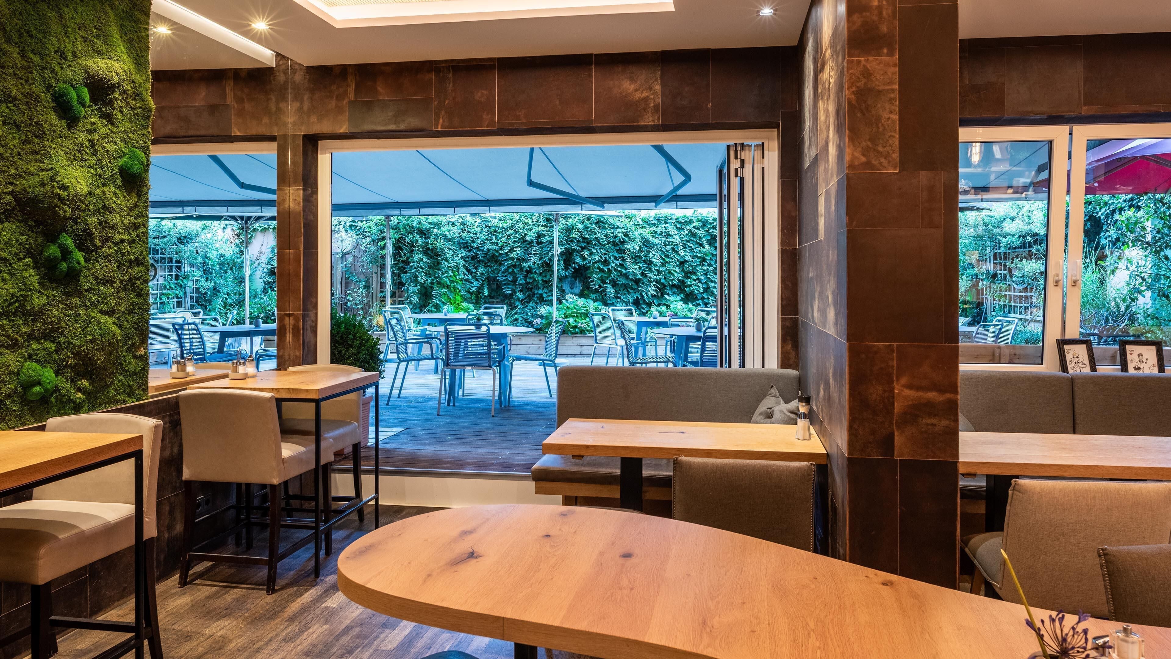 Un comedor interior con mesas y sillas de madera, una pared verde con musgo, y vistas del patio exterior.