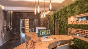 A modern reception desk with a computer monitor displaying a welcome page, a wooden table with chairs, and a green moss wall. - Bavaria Boutique Hotel (Munich)