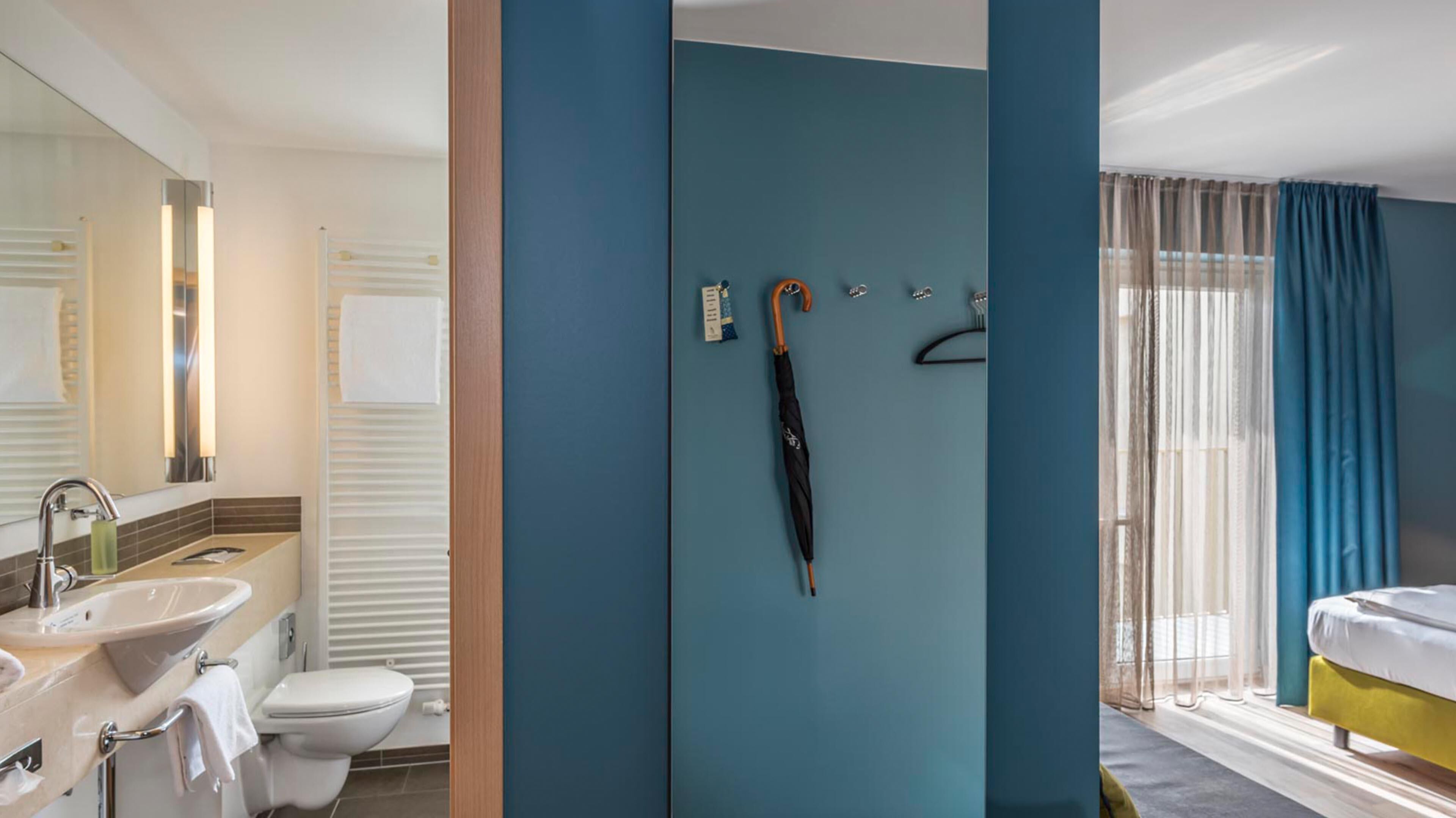 A bathroom with a sink, toilet, and mirror, a shower area with a towel rack, and a bed with a window.