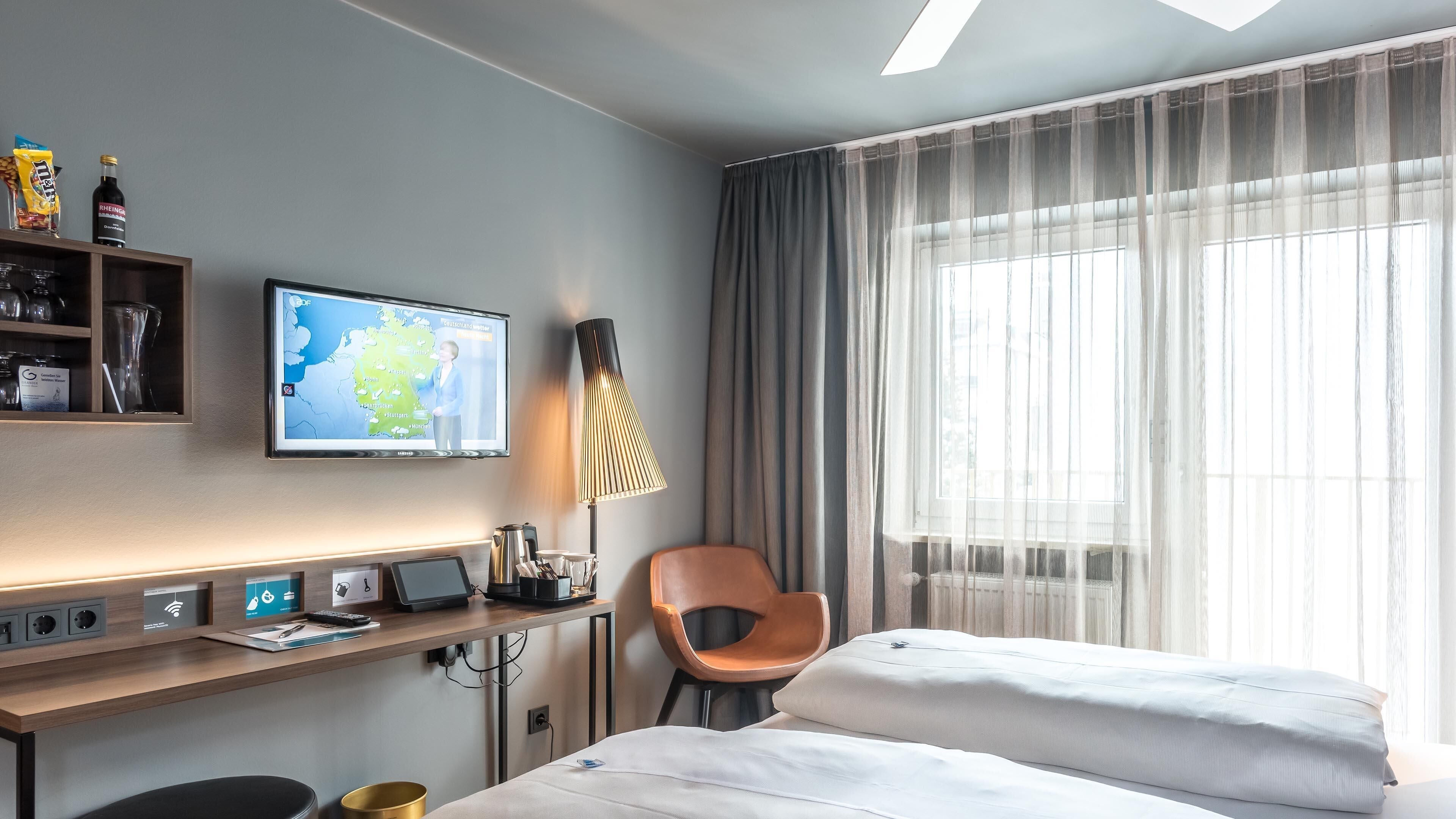 A hotel room with a bed, a desk with a computer, a TV displaying a map, a lamp, and a chair.