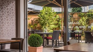 An outdoor patio area with wooden tables and chairs, potted plants, and umbrellas providing shade. - Bavaria Boutique Hotel (Munich)