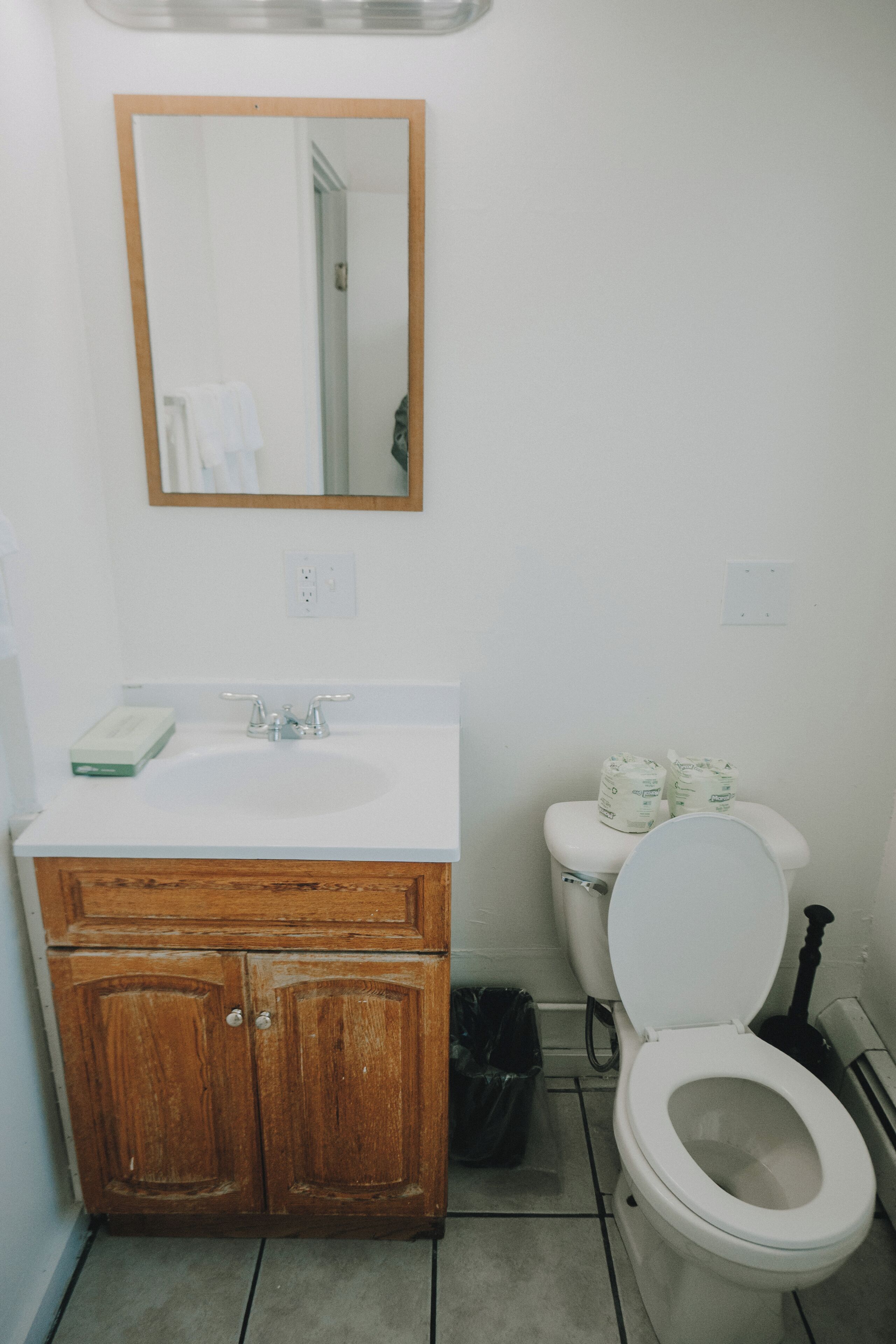 A bathroom with a wooden vanity, a white sink, a mirror, a toilet, and a trash bin.