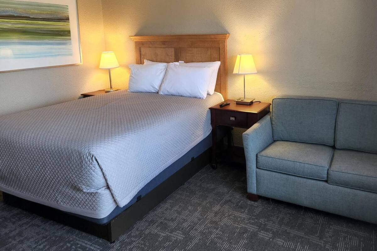 A hotel room with a large bed, two bedside tables with lamps, a gray armchair, and a painting on the wall.