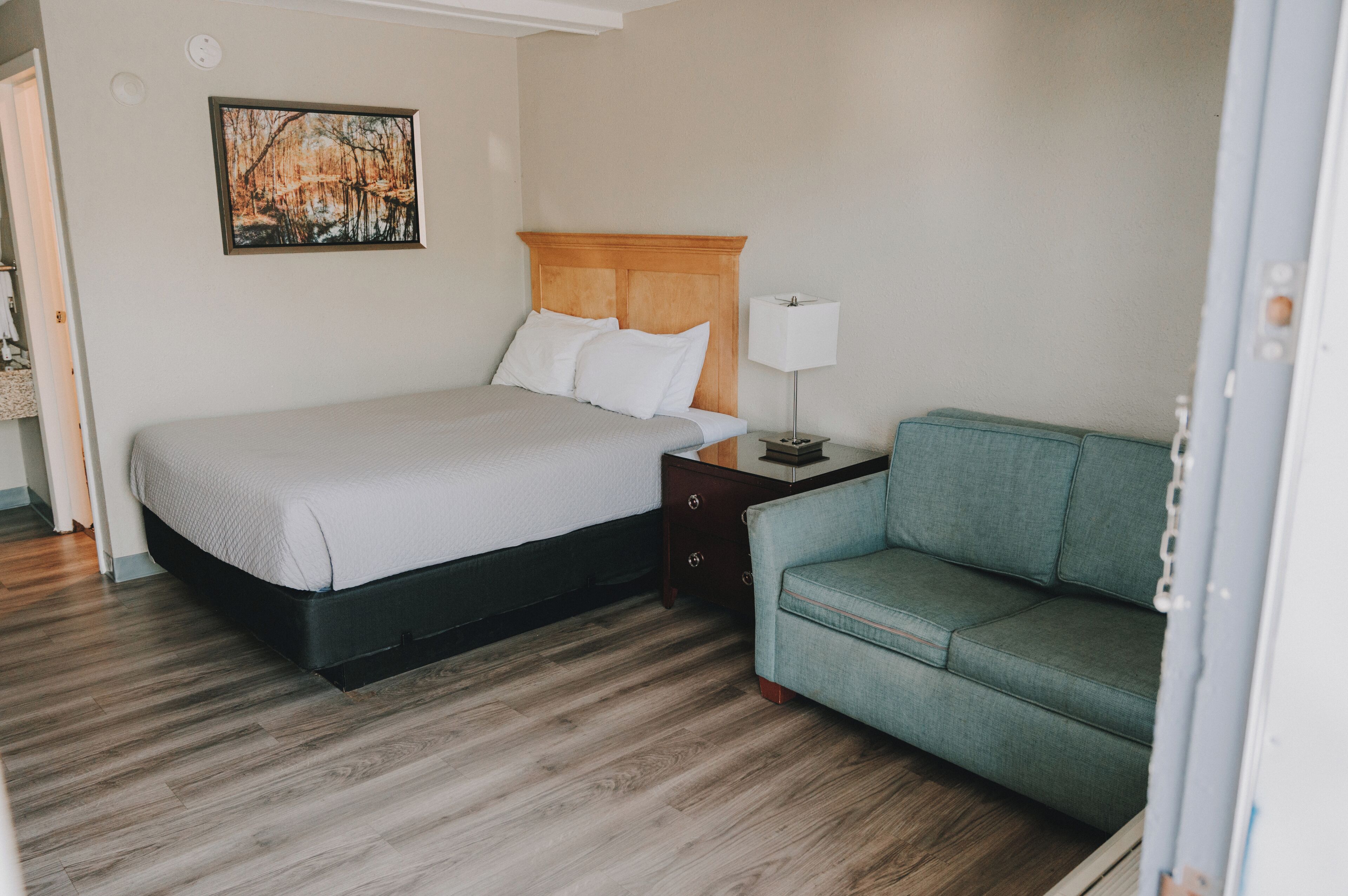Une chambre d’hôtel avec un lit, une table de chevet, une lampe, un fauteuil vert et un tableau au mur.