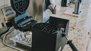 A waffle maker on a kitchen counter with a waffle sign, a coffee machine, and a bread display in the background.