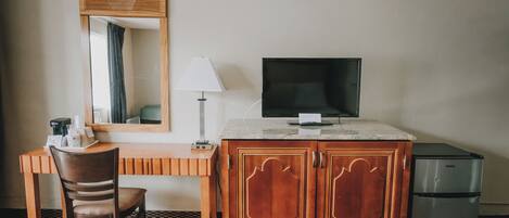 A hotel room with a wooden desk, a chair, a television, a cabinet, and a refrigerator.