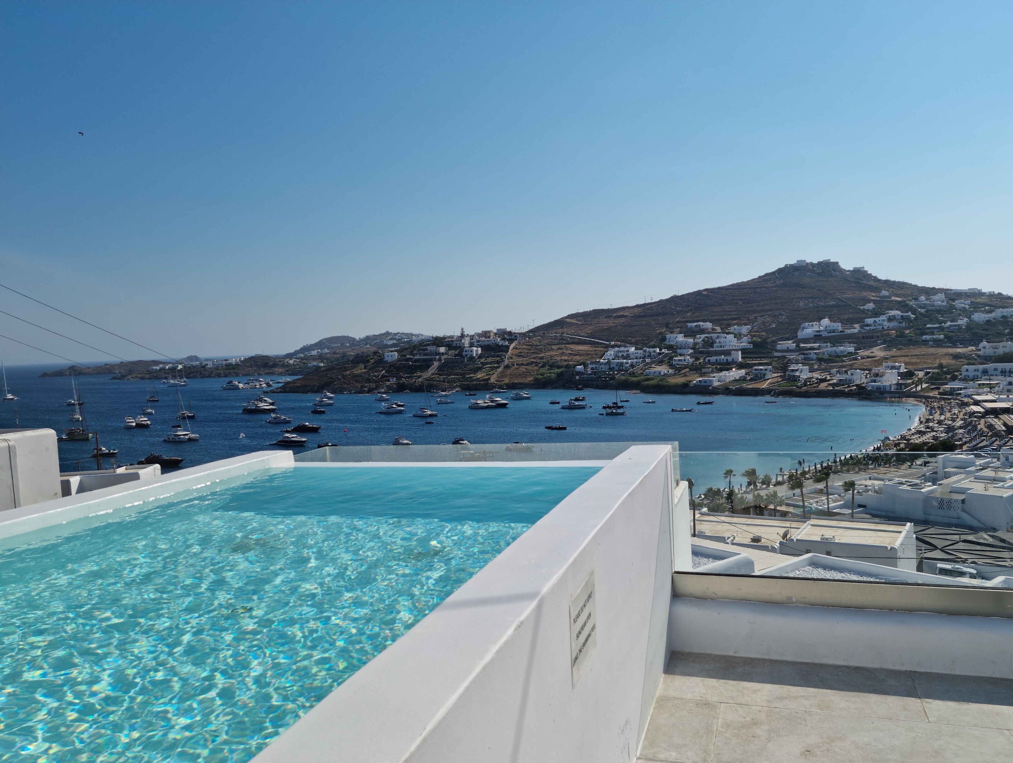 A clear blue pool with a view of boats on the sea and a hillside with white buildings.