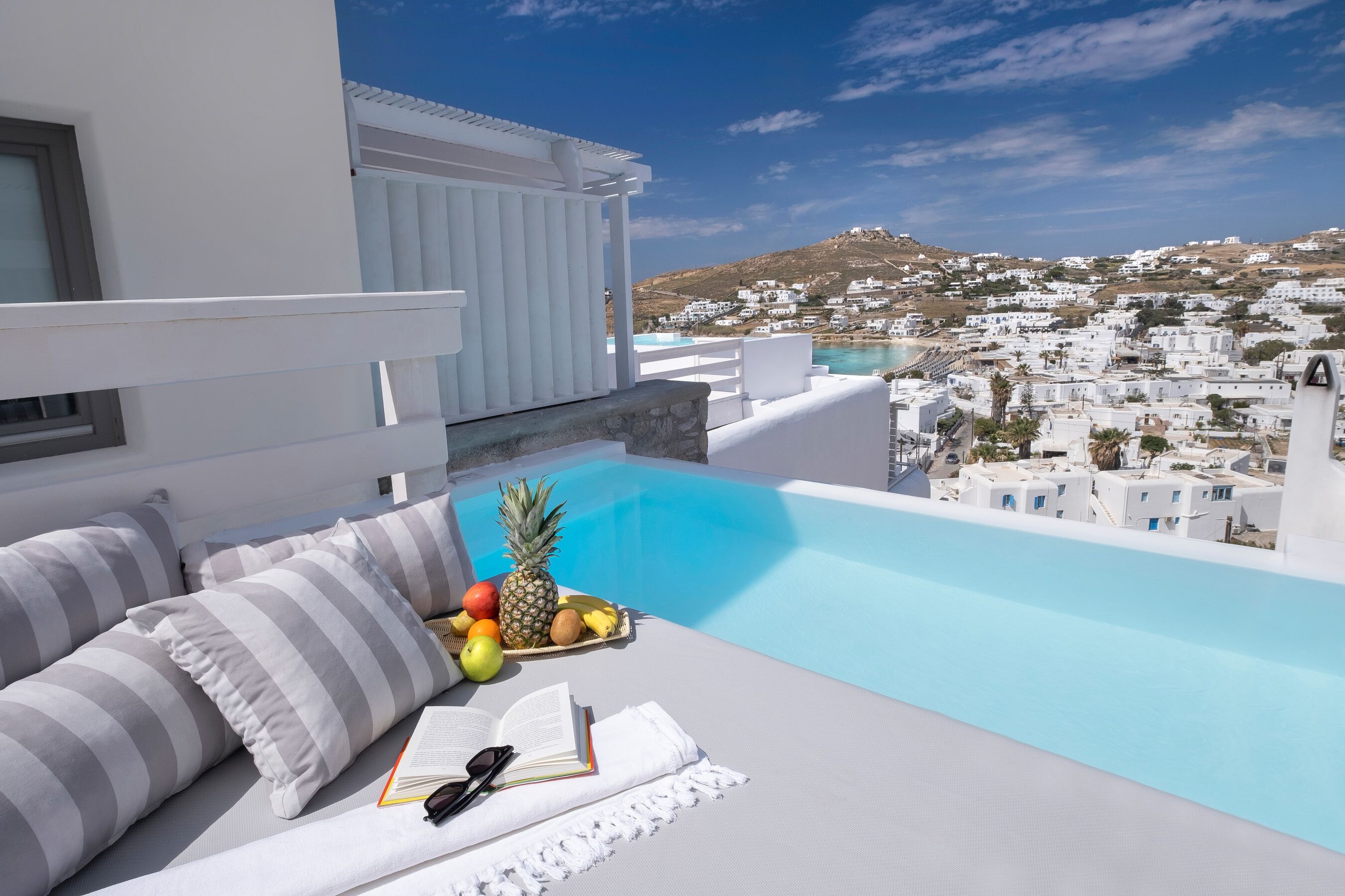 A rooftop lounge with a pool, white furniture, and a view of a white-washed town.