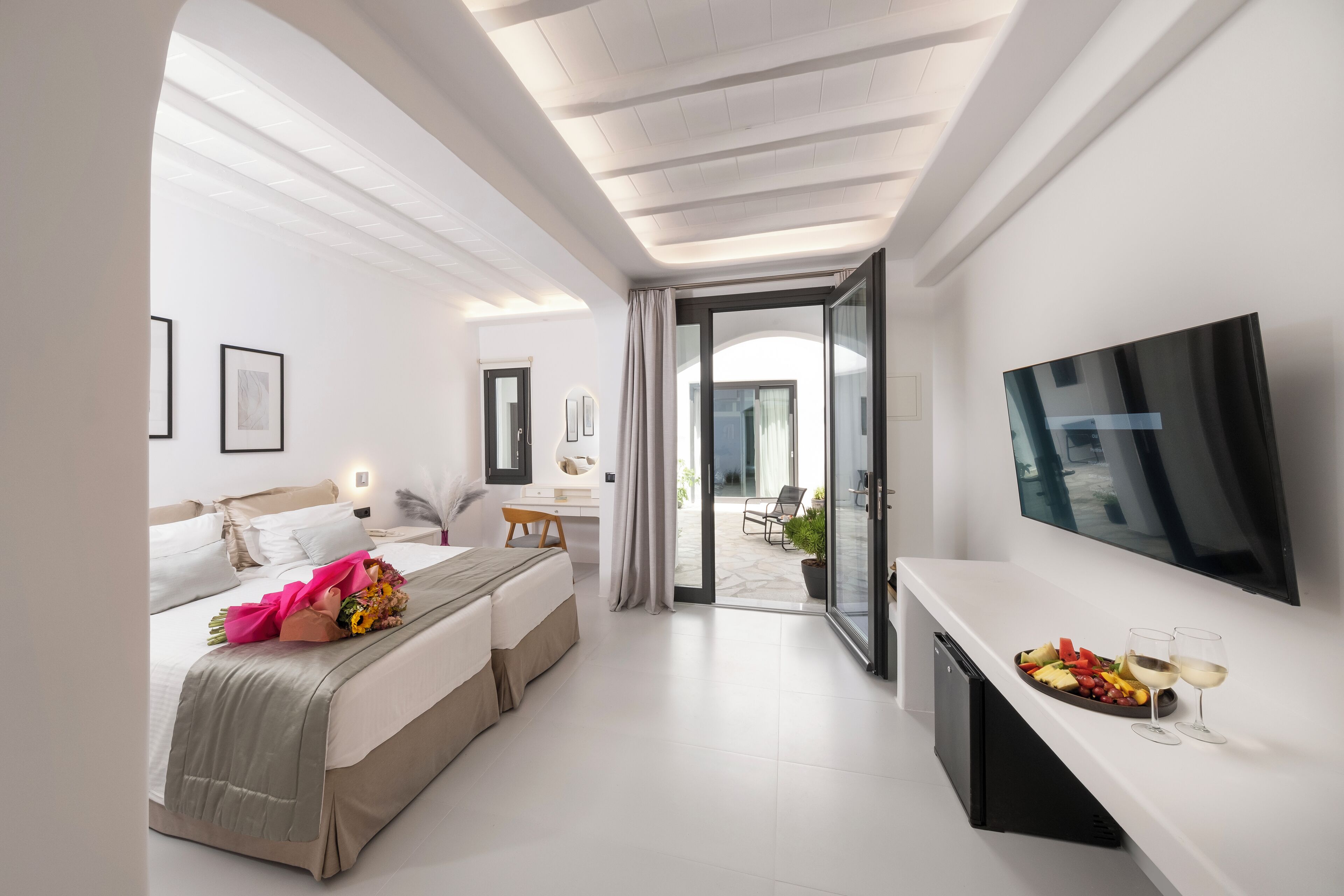 Una habitación de hotel moderna con cama, televisor, zona de comedor con bandeja de fruta y copas de vino, y balcón con mobiliario exterior.