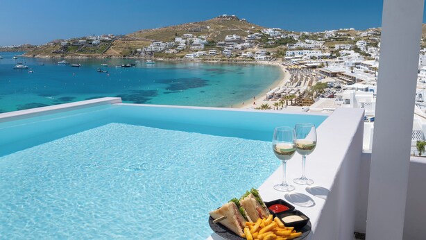 Una piscina con vistas a un pueblo costero, una mesa con comida y bebidas, y un cielo azul despejado.