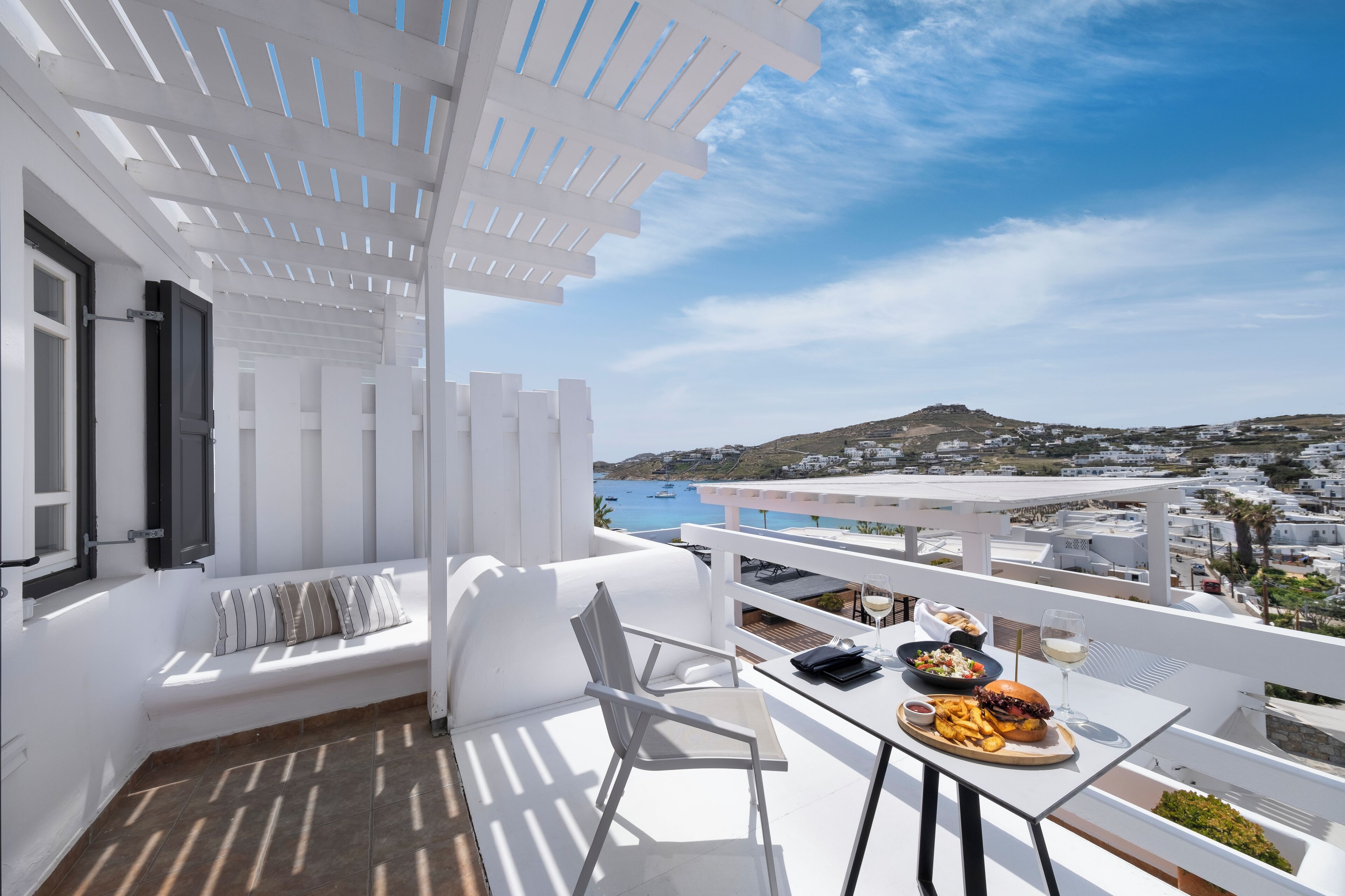 A balcony with a white table set for dining, a white sofa, and a view of the sea and hills.