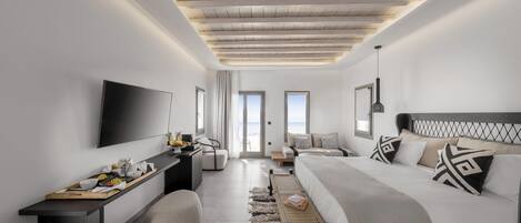 Una habitación de hotel moderna con una cama grande, un escritorio con televisor de pantalla plana, una zona de estar con vista al mar y un techo con vigas a la vista.