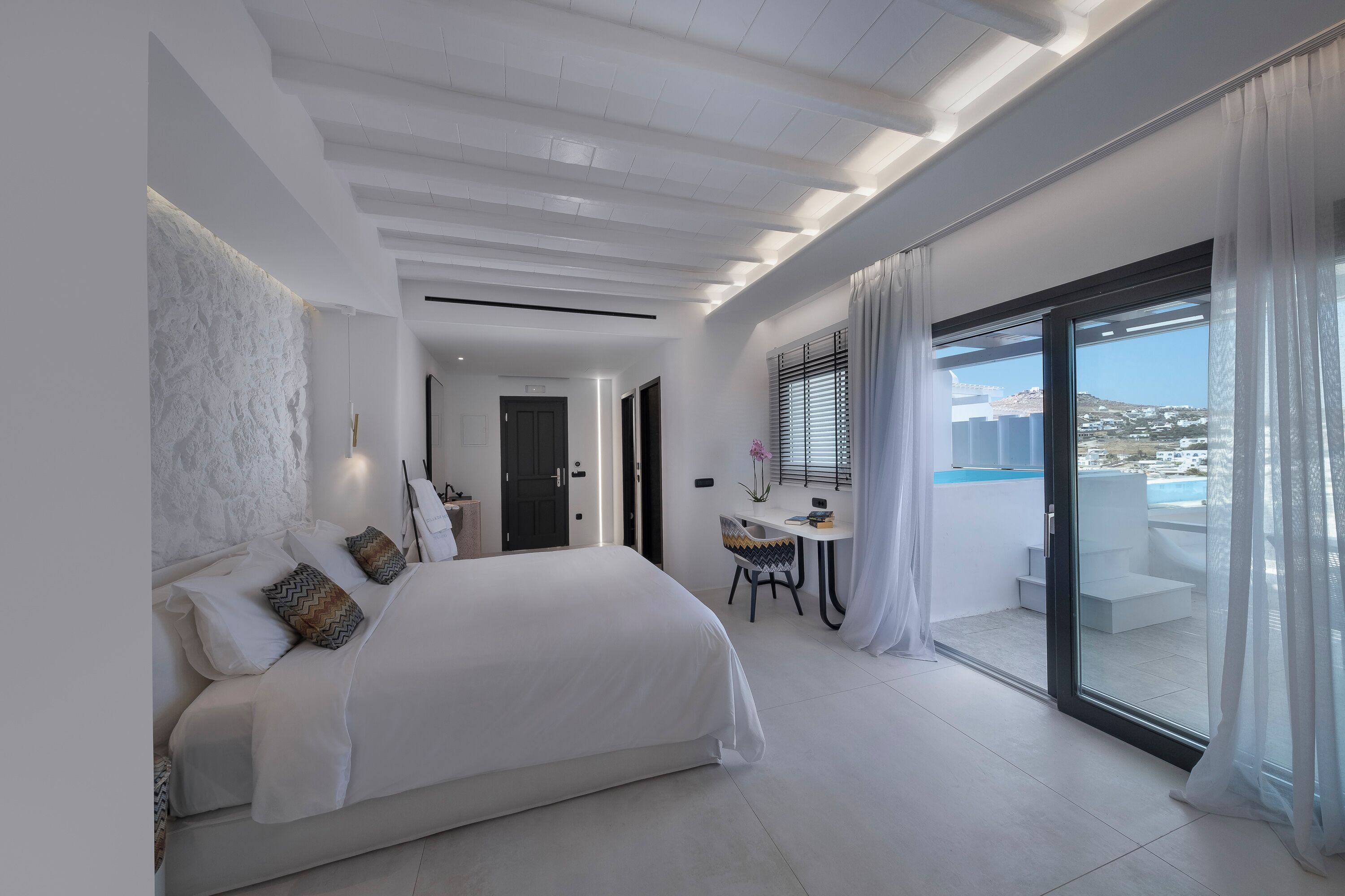 A modern bedroom with a large bed, a desk, and a balcony with a city view.