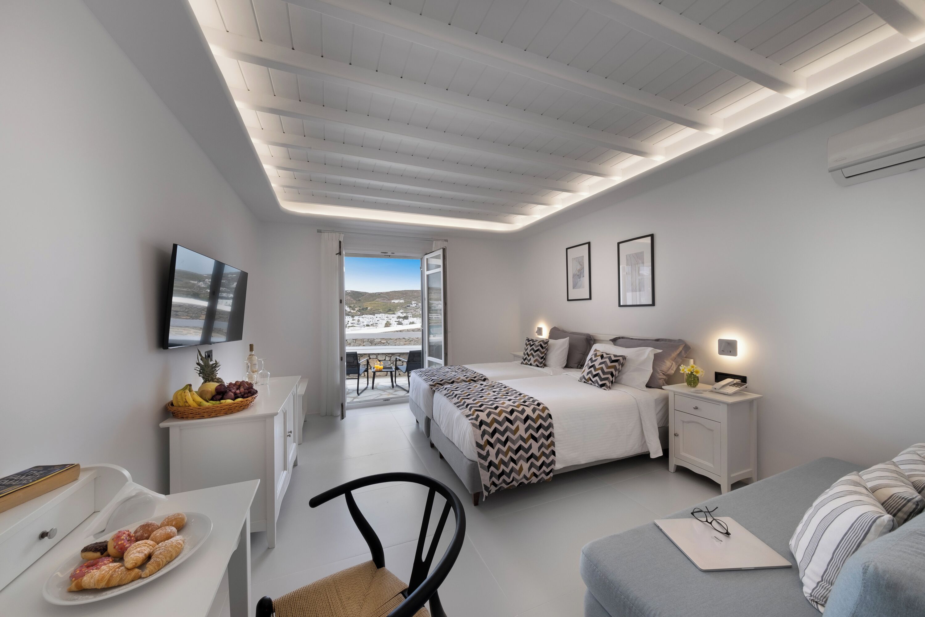 A modern bedroom with a bed, bedside tables, a desk with pastries, a chair, and a view through an open door.